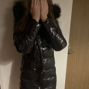 Svart glansig dunjacka med pälshuva - Säljer en snygg svart glansig dunjacka med pälshuva. Perfekt för kalla vinterdagar! Jackan har en dragkedja framtill och långa ärmar. Den är i mycket bra skick och håller dig varm och stilren hela säsongen. Den är aldrig använd. Pris kan även diskuteras!