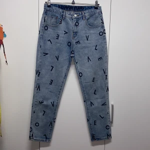 Blå jeans med bokstavsmönster som nya - Säljer ett par unika blå jeans med coolt bokstavsmönster över hela byxorna. De har en normal passform och är i bra skick. På baksidan finns det en liten reva som ger dem en extra edge. Perfekta för en avslappnad stil!