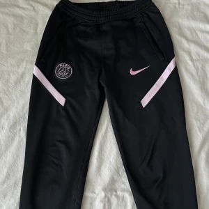 Svarta träningsbyxor från Nike x PSG - Säljer ett par svarta träningsbyxor från Nike- PSG. Byxorna har rosa detaljer och PSG-logga på ena benet. Perfekta för träning eller vardagsbruk. De har en elastisk midja och är gjorda i ett bekvämt material. Använd fåtal gånger, fortf mkt bra skick!!