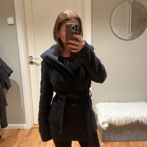 Mörkgrå kappa - Säljer en snygg mörkgrå kappa i mycket bra skick. Den har långa ärmar och ett bälte i midjan för en fin passform. Perfekt för höst och vinter, och den är både stilren och varm. Passar till både vardag och fest! Storleken är M men passar mig som bär XS och S💓💓