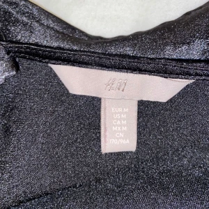 Svart glittrig topp från H&M - Säljer en snygg svart glittrig topp från H&M i storlek M. Toppen har en åtsittande passform och är perfekt för festliga tillfällen. Den har en elegant rynkad detalj på baksidan som ger en extra touch. Perfekt för att glittra på festen! ✨
