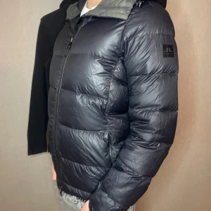 Svart pufferjacka från J.Lindeberg - Säljer en snygg svart pufferjacka från J.Lindeberg i mycket bra skick. Jackan har en dragkedja framtill och en praktisk huva. Perfekt för kalla höst- och vinterdagar. Den är både stilren och varm, vilket gör den till ett måste i garderoben!