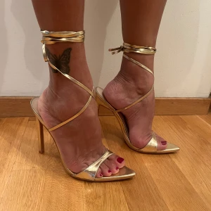 Guldiga klackskor med remmar - Snygga guldiga klackskor med spetsig tå och långa snören som knyts runt foten, dom är cirka 10 cm i klacken. Klackarna är från prettylittlething och är helt oanvända🤍