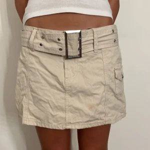 Beige cargokjol med stort bälte - Säljer en beige kortkjol med ett stort, coolt bälte och fickdetalj. Kjolen har ett brännmärke på baksidan, men annars i bra skick. Perfekt för sommardagar eller en avslappnad utekväll. Passar bra med en enkel topp och sneakers.