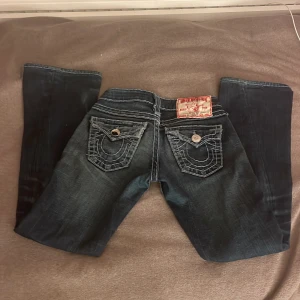 Mörkblå jeans från True Religion - Snygga mörkblå jeans från True Religion med ikonisk broderad design på bakfickorna. Jeansen har en lågmidjad passform och klassisk femficksdesign med knappar och dragkedja framtill. En av knappen e lite sönder framme men den funkar vara att detalijen är av annars är det utmärkt skick❤️ det är väldigt låg midja de och de är flare/bootcut