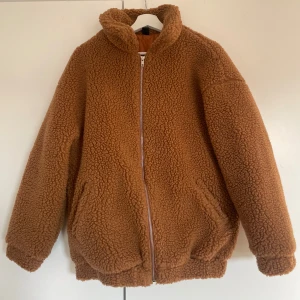 Brun teddyjacka med dragkedja - Mysig brun teddyjacka med dragkedja framtill. Jackan har en fluffig textur och är perfekt för kyligare dagar. Den har långa ärmar och en bekväm passform. Aldrig använd.