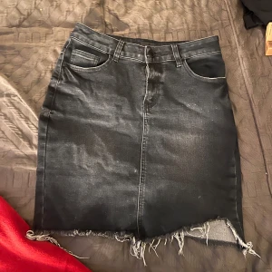 Svart jeanskjol med fransar - Snygg svart jeanskjol med fransig nederkant och slitna detaljer på bakfickorna. Kjolen har en klassisk femficksdesign och en bekväm passform. Perfekt för en avslappnad stil.