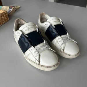 Valentino Garavani sneakers i vitt och blått - Snygga Valentino Garavani sneakers i vitt med blå detaljer. Skorna har en stilren design med snörning och den ikoniska rockstud-detaljen på hälen. Använda men fortfarande i väldigt gott skick. Släng iväg ett medellande om du har några frågor!