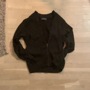 Svart ribbad kofta från Pierre Hector - Snygg svart kofta från Pierre Hector med ribbad design och dragkedja framtill. Perfekt för kyligare dagar och enkel att matcha med olika outfits.