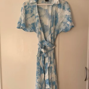 Blå och vit omlottklänning - Säljer en somrig blå och vit omlottklänning med korta ärmar och knytband i midjan. Klänningen är använd ett fåtal gånger men har en liten fläck i midjan som knappt syns. 