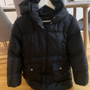 Svart dunjacka med huva - Säljer en svart dunjacka med huva och dragkedja. Jackan har långa ärmar och praktiska fickor med knappar framtill. Perfekt för kalla dagar.