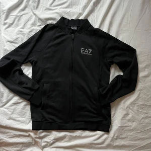 Svart tröja från Emporio Armani - Säljer en stilren svart jacka från Emporio Armani med dragkedja och EA7-logga både fram och bak. Jackan har långa ärmar och är tillverkad i ett syntetmaterial. Nypris 999:- SEK