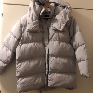 Grå pufferjacka från Boohoo - Säljer en snygg grå pufferjacka från Boohoo. Jackan har en dragkedja framtill och en stor huva för extra värme. Perfekt för kyliga dagar med sin vadderade design.