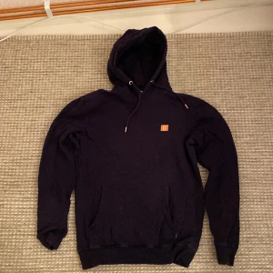 Svart hoodie från Les Deux - Säljer en stilren svart hoodie från Les Deux. Den har en klassisk design med en känguruficka och justerbar huva med snören. Perfekt för en avslappnad look.