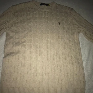 Beige stickad tröja från Ralph Lauren - Säljer en klassisk beige stickad tröja från Ralph Lauren med kabelstickat mönster och en broderad brun logotyp på bröstet. Tröjan har långa ärmar och ribbstickade muddar vid ärmslut och nederkant. Perfekt för en stilren look.