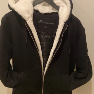 Svart jacka från Moose Knuckles - Säljer en svart jacka från Moose Knuckles med vit foder i huvan. Jackan har dragkedja och en snygg detalj på ärmen. Perfekt för kyligare dagar. Kvitto och Tags samt en Galje följer med vid köp. Köpt för 2 månader sedan på Mal of scandinavia. Hör av er vid mer funderingar }{ 