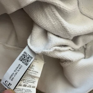 Vit hoodie från C.P. Company - Säljer en stilren vit hoodie från C.P. Company. Tröjan har en praktisk ficka framtill och en unik detalj med en lins på ärmen. Perfekt för en avslappnad stil.