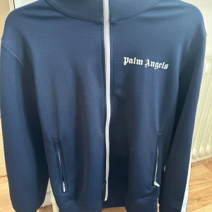 Mörkblå track jacket från Palm Angels - Säljer en snygg mörkblå track jacket från Palm Angels med vita detaljer. Jackan har dragkedja framtill och vita ränder längs ärmarna.
