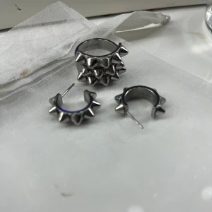 Silvriga örhängen och ring med nitar - Snygga silvriga örhängen och matchande ring med nitar. Perfekt för en edgy stil. Örhängena är små hoops med spetsiga nitar och ringen har en rad av nitar runt om. Ett coolt set för att sticka ut! Från Edblad! Ring och örhängen säljs tillsammans för 450!