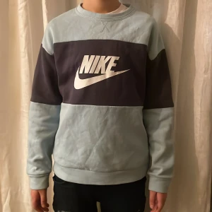 Blå och mörkgrå sweatshirt från Nike - Säljer en snygg blå och mörkgrå sweatshirt från Nike. Tröjan har ett stort Nike-logotyptryck på framsidan och är långärmad med ribbade muddar. Perfekt för en avslappnad stil.