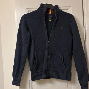 Mörkblå zip tröja från Polo Ralph Lauren - Säljer en snygg mörkblå kofta från Polo Ralph Lauren med dragkedja och broderad röd logga på bröstet. Koftan har ribbade muddar och en randig krage i rött och gult. Perfekt för kyliga dagar! Skick 7,5/10 