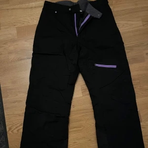 Svarta överdragsbyxor - Säljer ett par svarta överdragsbyxor med lila dragkedjor och justerbart bälte. Perfekta för vinterns kalla dagar. Byxorna har en praktisk ficka med dragkedja på sidan. Skriv om du har fler frågor eller vill se fler bilder💕(pris går att diskutera)