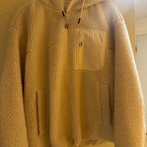 Beige teddyjacka med huva - Mysig beige teddyjacka med dragkedja och huva. Jackan har en praktisk bröstficka med dragkedja och två sidofickor. Perfekt för kyliga dagar när du vill hålla dig varm och bekväm.