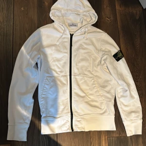 Vit hoodie från Stone Island - Säljer en stilren vit hoodie från Stone Island med dragkedja och huva. Tröjan har en svart logga på ärmen och praktiska fickor framtill. Perfekt för en avslappnad stil. Storlek s