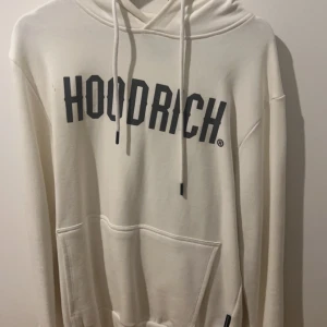 Vit hoodie från Hoodrich - Säljer en stilren vit hoodie från Hoodrich med svart text på framsidan. Den har en klassisk känguruficka och justerbar huva med dragsko. Perfekt för en avslappnad stil. Väldigt fin skick på hoodien