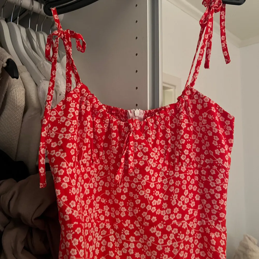 Charmig röd klänning med vita blommor och knytband vid axlarna. Perfekt för sommarens varma dagar. Den har en smockad överdel som ger en bekväm passform och en luftig kjol.. Mekot.