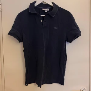 Mörkblå pikétröja från Lacoste - Säljer en stilren mörkblå pikétröja från Lacoste. Tröjan har korta ärmar och en klassisk krage med knappar. 