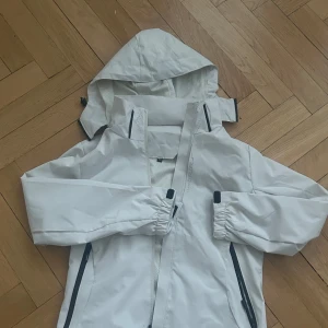 Skidjacka - Säljer en vit skidjacka i helt nytt skick med justerbar huva och dragkedja. Jackan har fickor med dragkedja. Funkar även som skid och vindjacka.