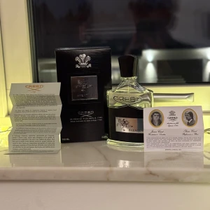 Creed Aventus Parfym - Exklusiv Creed Aventus parfym i en elegant glasflaska med svart etikett och silverdetaljer. Förpackningen är stilren och svart med Creed-logotypen i silver. Flaskan rymmer 100 ml av denna ikoniska doft, känd för sina fruktiga och träiga noter.