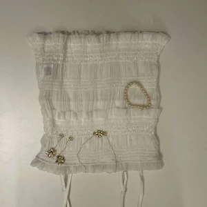 Vit smockad tubtopp - Säljer en söt vit tubtopp med smockad design och subtila glittriga detaljer. Toppen är axelbandslös och har en romantisk känsla med sina rynkade partier. Perfekt för en somrig look eller en speciell kväll.