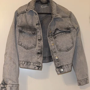 Grå jeansjacka - Snygg grå jeansjacka med en vintage look. Jackan har en croppad passform och knappar framtill. Två bröstfickor med lock och långa ärmar med knappdetaljer vid ärmsluten. Perfekt för en avslappnad stil.