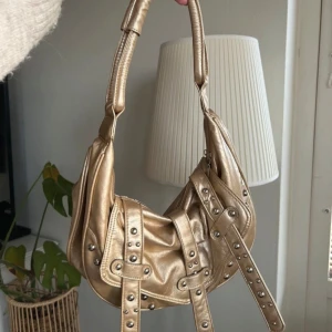Guldig axelväska med nitar - Snygg guldig axelväska med coola nitdetaljer och dragkedja. Perfekt för att ge din outfit en extra edge. Väskan har en unik design med flera remmar och en bekväm axelrem! Köpt secondhand utomlands ⭐️