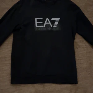 Emporio Armani tröja - Snygg svart tröja från Emporio Armani med EA7-logga på bröstet. Perfekt för en stilren look. Tröjan har långa ärmar och en klassisk passform. Tröjan är i storlek M men passar även S