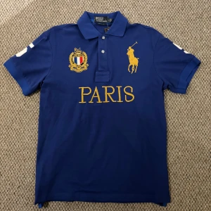 Ralph Lauren chief keef polo - Säljer en snygg blå pikétröja från Ralph Lauren med gul broderad logga och texten 'PARIS' på framsidan. Tröjan har en klassisk krage och knappar framtill. På baksidan finns en stor vit siffra 5. 