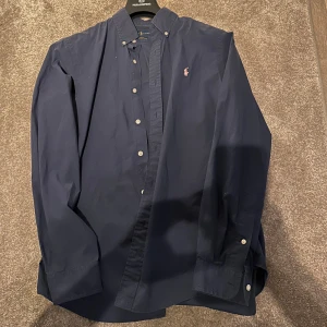 Mörkblå skjorta från Ralph Lauren - Snygg mörkblå skjorta från Ralph Lauren i slim fit. Tillverkad i 100% bomull med stretch för extra komfort. Skjortan har långa ärmar och knappar framtill samt en broderad logga på bröstet.