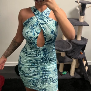 Turkos mönstrad klänning - Snygg turkos klänning med ett abstrakt mönster i olika blå nyanser. Klänningen har en unik design med en cut-out framtill och knytning i ryggen. Den är ärmlös och har en tight passform som framhäver figuren. Perfekt för en kväll ute!