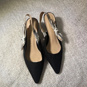 Svarta slingback pumps från Christian Dior - Eleganta svarta slingback pumps från Christian Dior med spetsig tå och dekorativa band med logotyp. Perfekta för en stilren look.