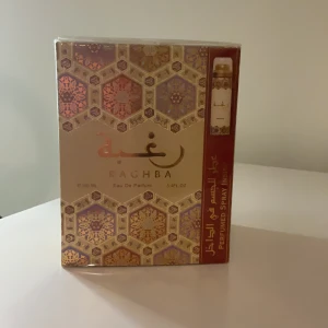 Lattafa Raghba Eau De Parfum 2 I 1 set. - Raghba Eau De Parfum i en elegant förpackning med ett intrikat mönster i guld och lila toner. Flaskan innehåller 100 ml av en rik och exotisk doft som ger en känsla av lyx och mystik.  Den är 2 i 1 set av 100 ml parfym + body spray. Perfekt för den som vill ha en unik och minnesvärd doftupplevelse. Oöppnat! Plast är kvar!