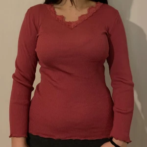 Röd ribbad topp med spetsdetaljer - Säljer en snygg röd ribbad topp med spetsdetaljer vid halsringningen. Toppen har långa ärmar och en figurnära passform. Perfekt för att lägga till en touch av stil i din garderob.