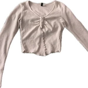 Beige ribbad topp - Säljer en beige ribbad topp med långa ärmar och en snygg rynkad detalj framtill. Toppen är croppad och har en v-ringad design som ger en trendig look. Perfekt för att styla med dina favoritjeans eller kjol.