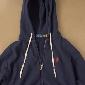 Mörkblå hoodie från Ralph Lauren - Säljer en snygg mörkblå hoodie från Ralph Lauren med dragkedja och huva.(I storlek XS)Den har en klassisk design med det ikoniska röda logotypbroderiet på bröstet.