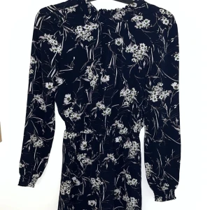 Svart blommig klänning - Säljer en elegant svart klänning med vita blommor som går under knäna. Klänningen har långa ärmar och en smickrande passform med elastisk midja. Perfekt för en somrig och fin look. 