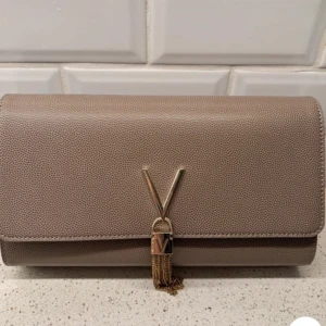 Beige kuvertväska med kedja - Snygg beige kuvertväska med guldfärgad kedja och dekorativ tofsdetalj. Väskan har en elegant V-formad metallapplikation på framsidan. Perfekt för att ge en stilren touch till din outfit.