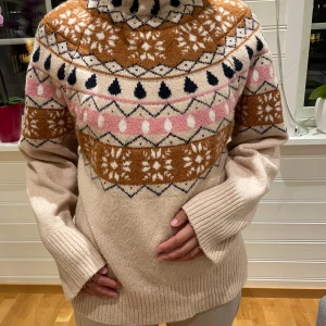 Beige stickad tröja med mönster - Säljer en mysig beige stickad tröja med ett färgglatt mönster i brunt, rosa och svart. Tröjan har långa ärmar och en hög krage, perfekt för kyliga dagar. Den har ett traditionellt mönster som ger en retro känsla.