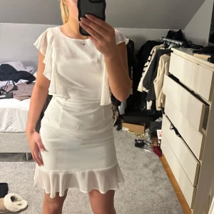 Vit klänning med volangdetaljer - Säljer en elegant vit klänning med volangdetaljer vid ärmarna och fållen. Klänningen har en dragkedja i ryggen och korta ärmar. Perfekt för studenten! Använd på min konfirmation en gång och köptes i Danmark! 🥰