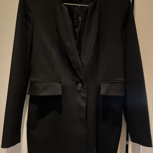 Svart kavaj med knapp - Elegant svart kavaj med en enkel knapp framtill. Den har en stilren design med långa ärmar och en klassisk passform. Perfekt för en sofistikerad look.